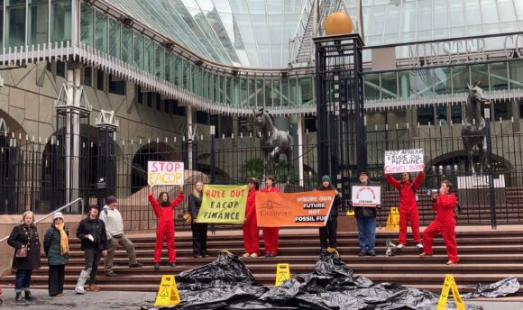 A group of activists dressed mostly in red jumpsuits stand in front of an office building with large glass windows. They are holding signs and banners with different phrases about "stopping EACOP" there are yellow signs on the ground and a black fake oil spill on the ground.